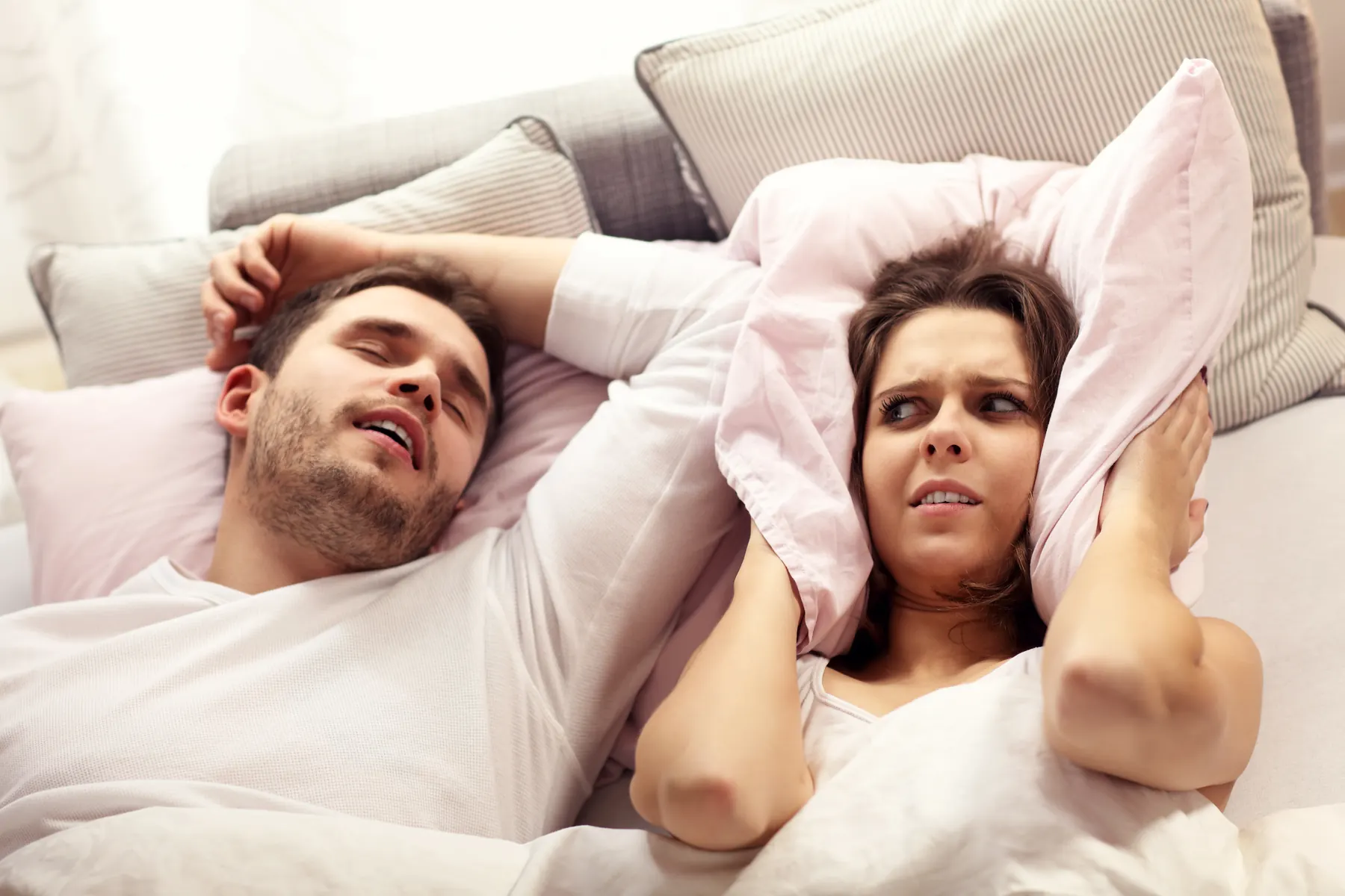 Couple in bed, one partner grinding or snoring while the other covers their ears